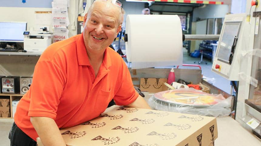 En glad man håller i en stor brun låda framför maskinen som minimerar emballaget. 