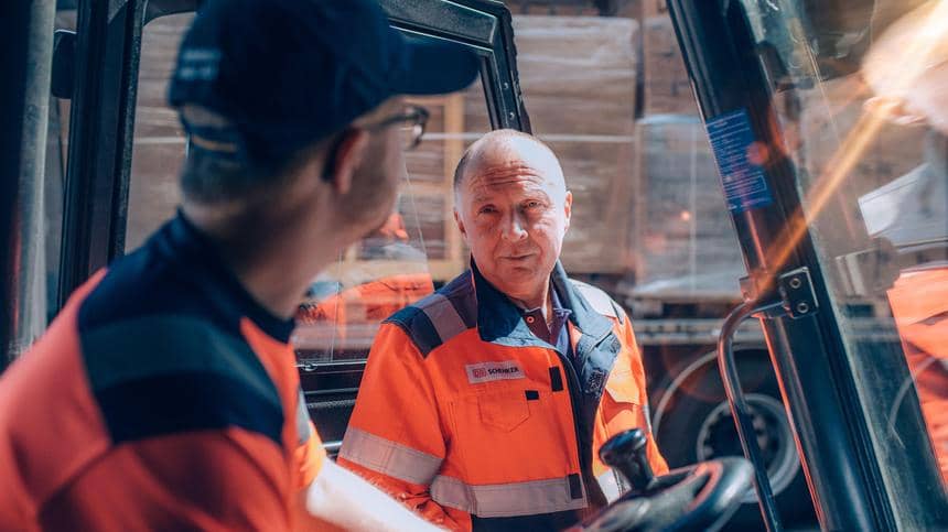 Två lastbilschaufförer i arbetskläder med DB Schenker-logo pratar intill lasttruckar. 