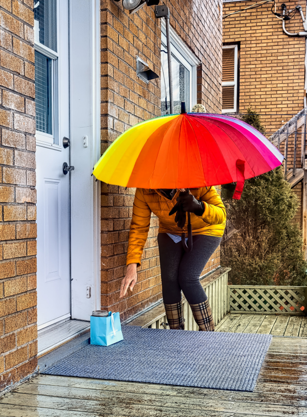En kvinna med paraply i regnbågens alla färger står vid en ytterdörr för att hämta ett paket som levererats framför ytterdörren. 