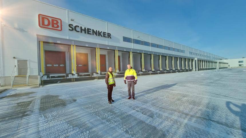 Två män i reflexjackor står på en snöig väg framför en stor lagerbyggnad med DB Schenker-logo. 