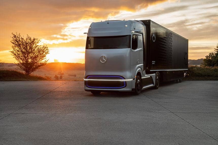 En stor grå-svart lastbil av märket Mercedes-Benz står på en asfalterad väg i ett öken med solnedgång i horisonten. 