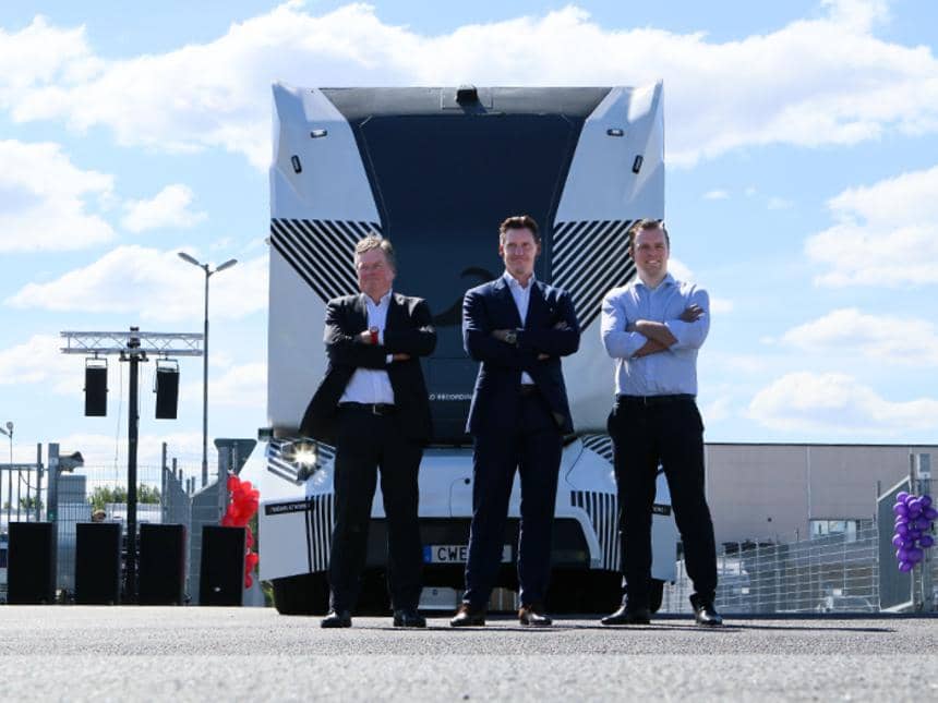 Tre män i kostym står med händerna korsade framför en stor självkörande lastbil en solig dag på terminalen. 