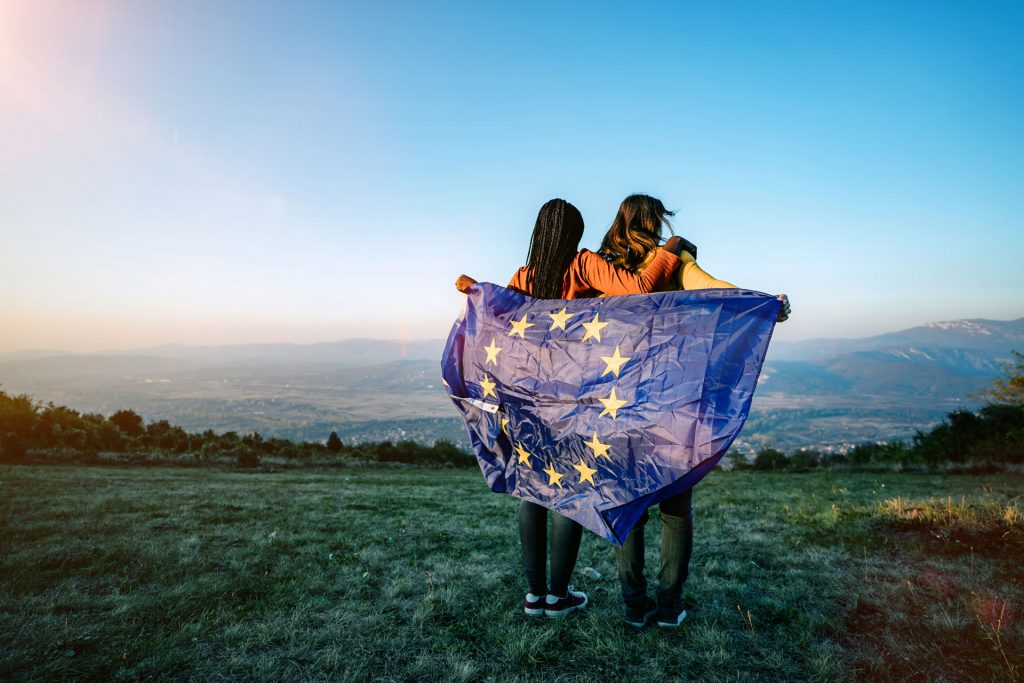 Två unga kvinnor står på ett grönt fält och håller en EU-flagga.