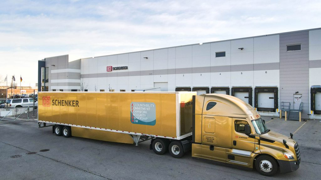 Guldfärgad lastbil plus släp som står utanför en terminal. På lastbilen och terminalen står det DB Schenker.