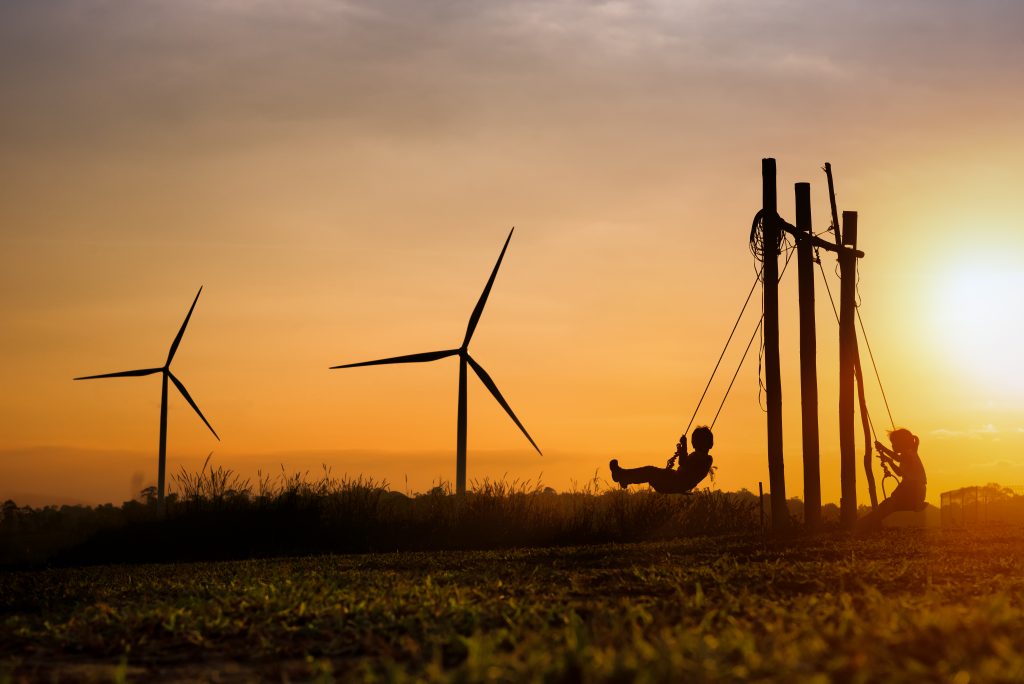 Två barn som gungar på ett fält i solnedgången. I bakgrunden syns två vindkraftverk.