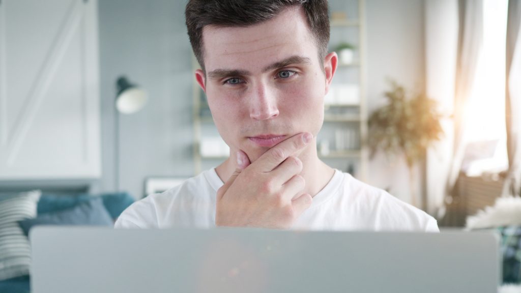 En ung man sitter framför sin laptop i ett ljust rum med en soffa, lampa och bokhylla i bakgrunden. Han ser fundersam ut och lutar hakan mellan pekfingret och tummen.