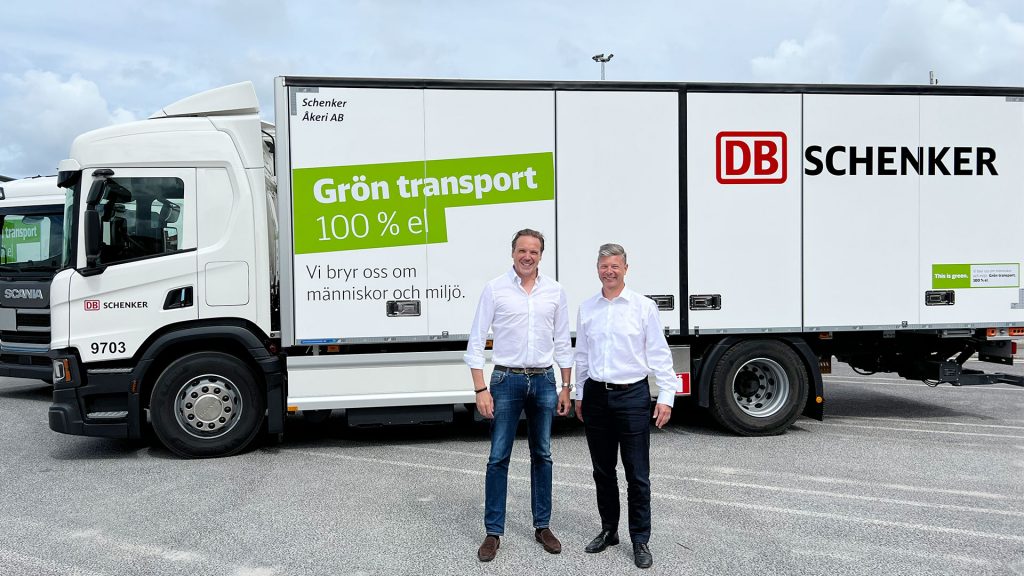 Två män står framför en ellastbil på en parkering. På lastbilen står det grön transport 100% el och DB Schenker.